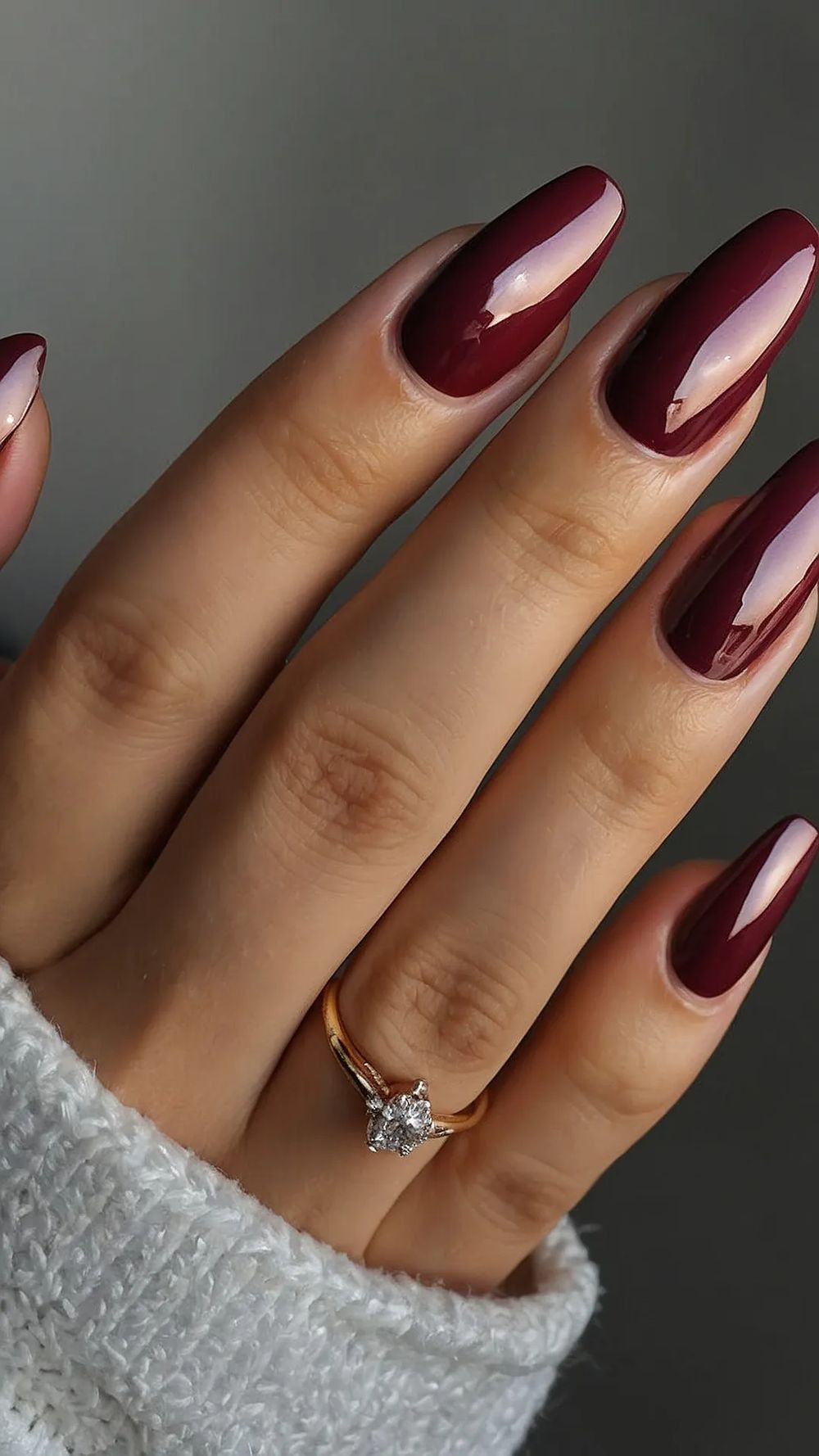Glossy burgundy almond nails with a high shine gel finish, hand tucked into a grey knit sleeve