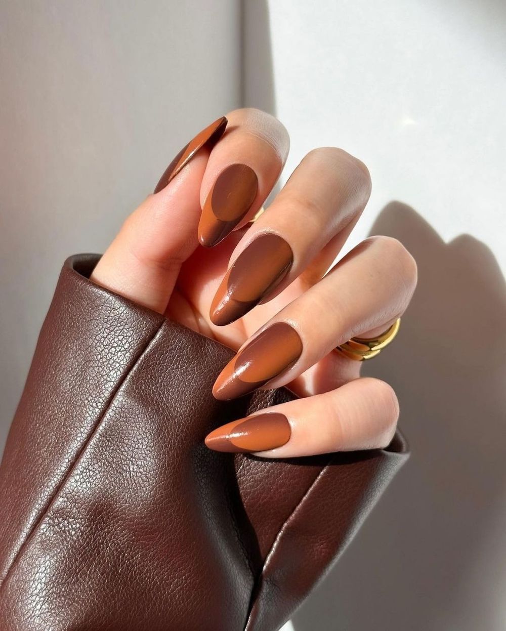Caramel ombre almond nails fading from chocolate to toffee, hand in a brown leather sleeve