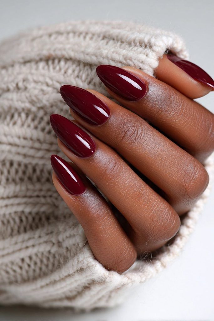 Glossy burgundy almond nails on dark skin, hand tucked into a cream knit sweater sleeve