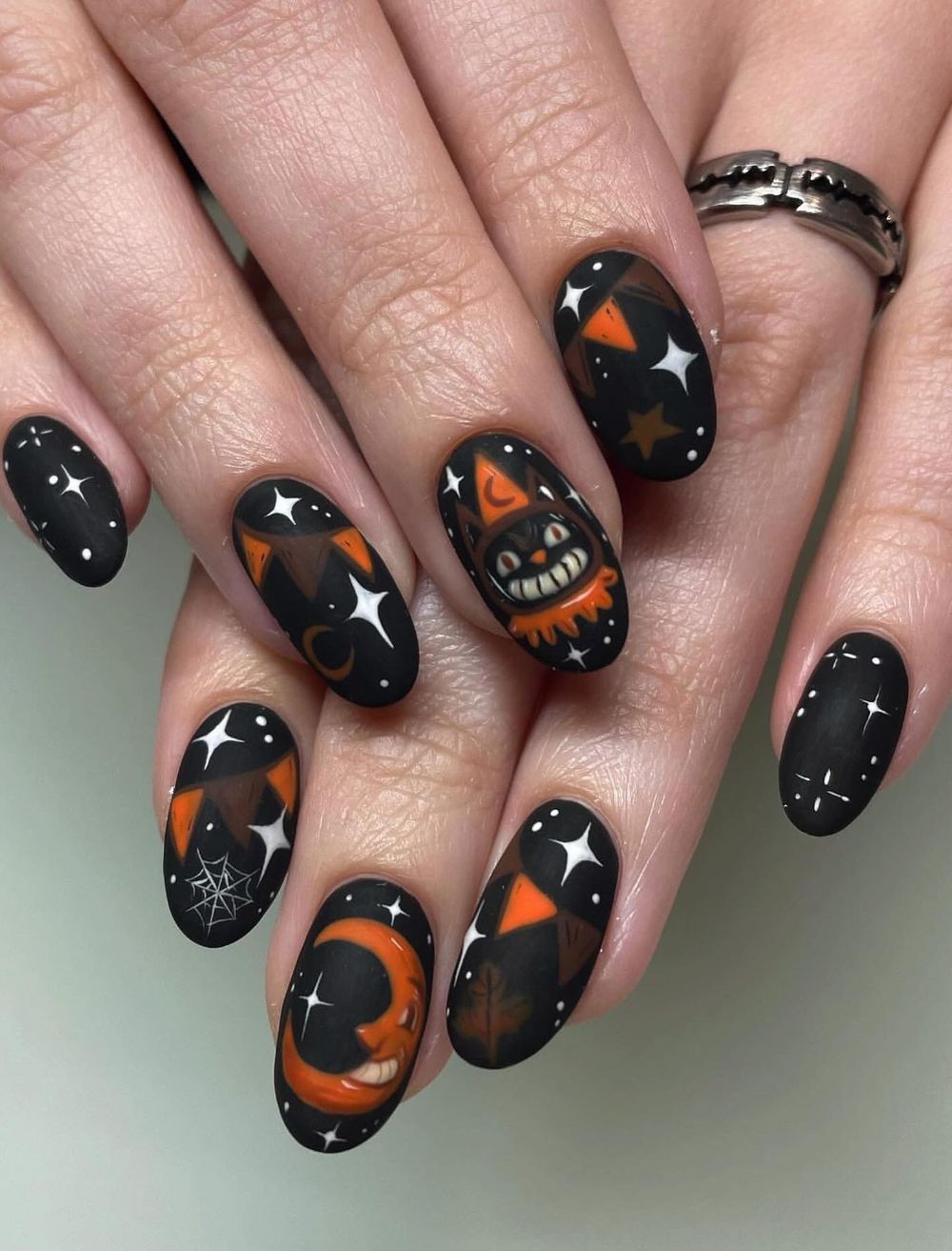 Black oval nails with orange jack-o-lantern faces, stars and tiny webs, glossy Halloween nail art on both hands