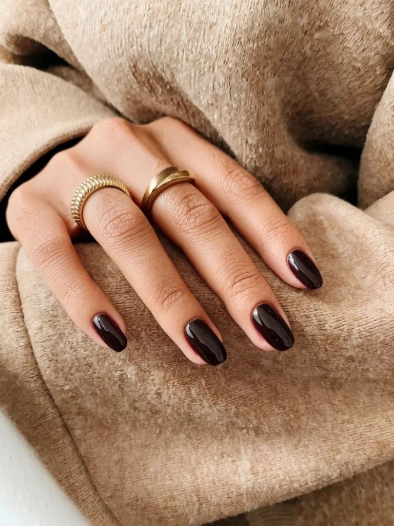 Short chocolate brown nails on a hand resting on a beige coat, gold rings, natural short length